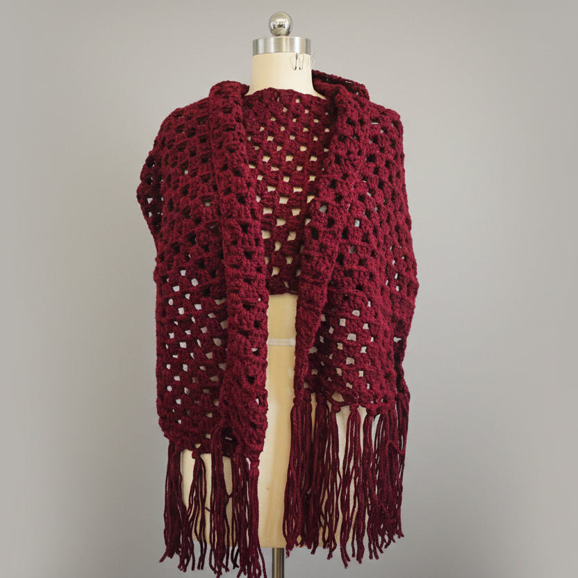 Maroon crocheted over sized wrap with fringe on a mannequin against a gray background