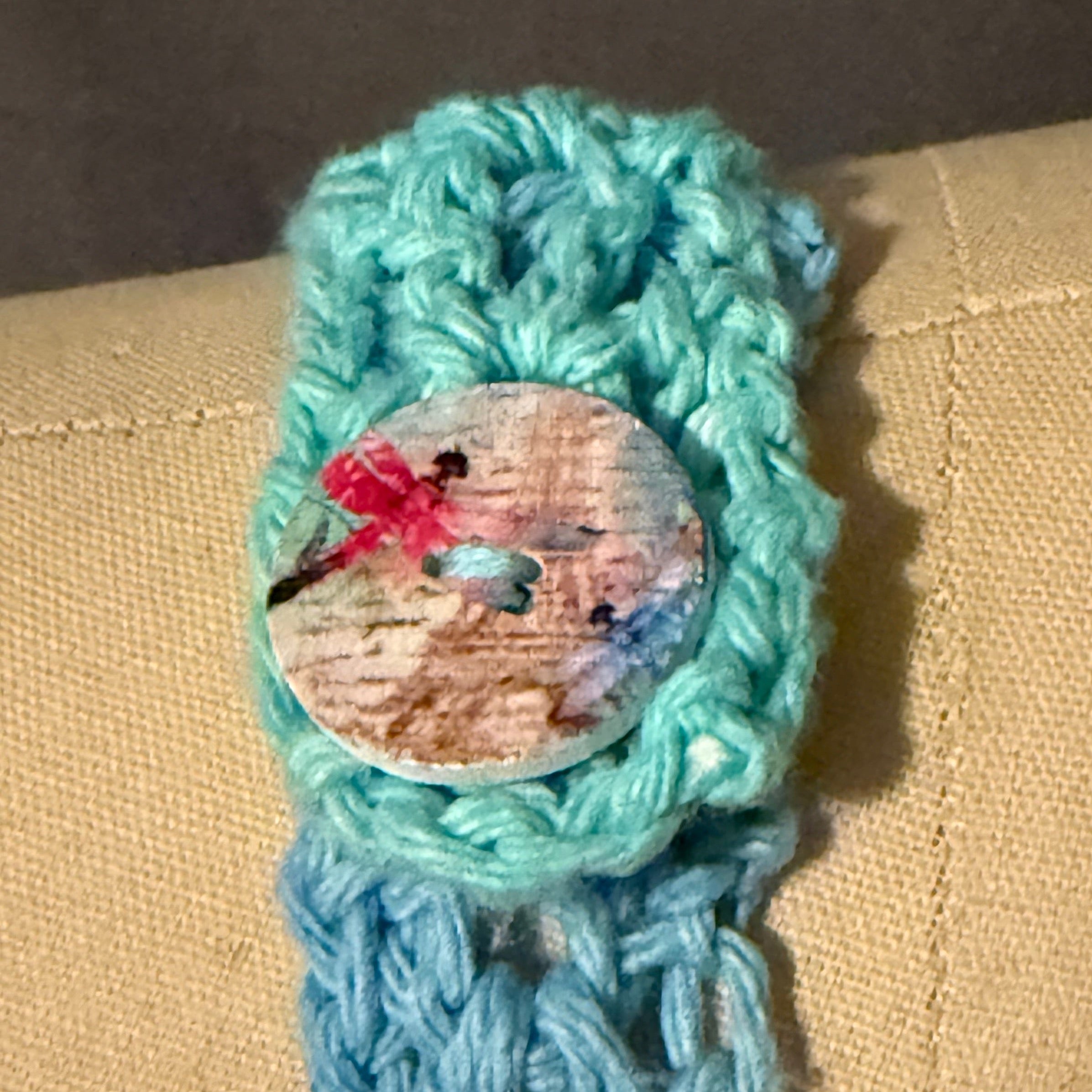 Crocheted blue item with a button featuring a dragonfly design on a beige surface.